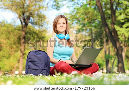 Female student with headphones relaxing in park and working on laptop