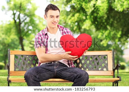 Smiling guy sitting on a bench and holding a red heart in park