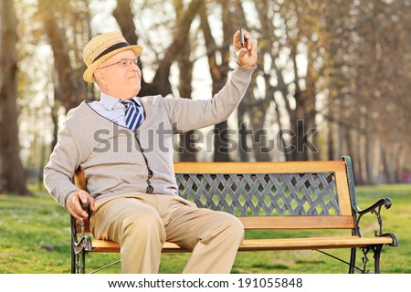 Elderly gentleman taking a selfie wit cell phone, seated on bench in park