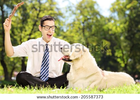 Guy with tie and glasses seated on a grass playing with labrador retriver dog in a park