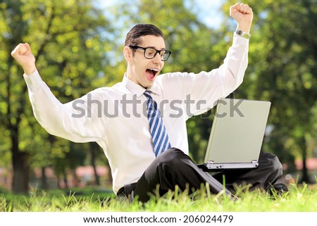 Young man cheering and watching TV on a laptop in park on a sunny day