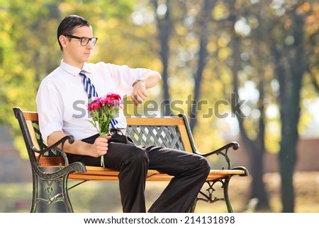 Young man holding flowers and checking the time seated on bench in park