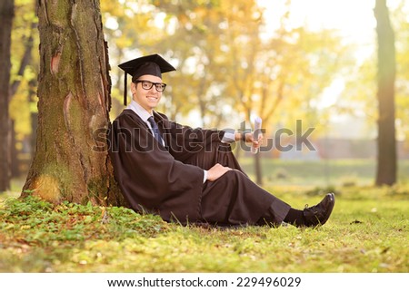 Cheerful college graduate sitting by a tree in a park