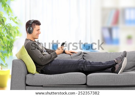 Young guy lying on sofa and listening to music on his cell phone isolated on white background