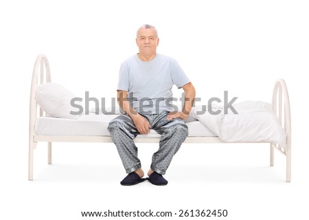 Senior man in pajamas sitting on a bed isolated on white background