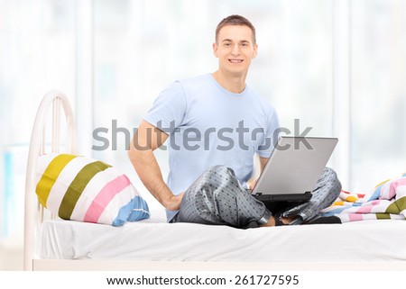 Man in pajamas working on a laptop seated on a bed at home