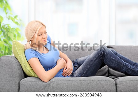 Blond woman with a painful expression sitting on a grey sofa at home with her hands placed on her stomach