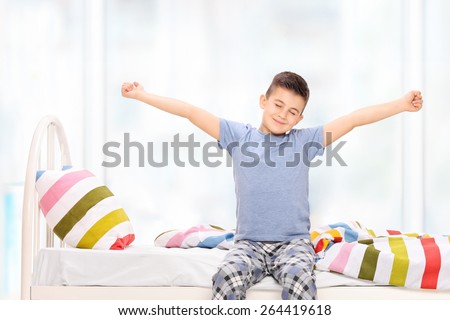 Sleepy little boy in pajamas stretching himself seated on a bed at home