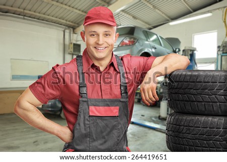 Cheerful male mechanic leaning on a stack of tires and posing in a garage with a car behind him