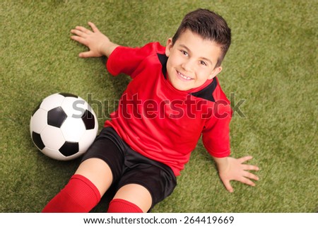 High angle shot of a junior soccer player in red jersey, sitting on a green field and looking at the camera, with a soccer ball beside him