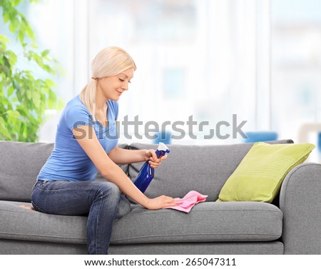 Young blond housewife cleaning a couch with a rag and a cleaning spray at home