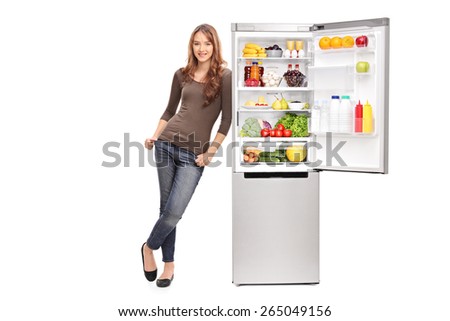 Full length portrait of a casual young girl leaning on an opened refrigerator full of food isolated on white background