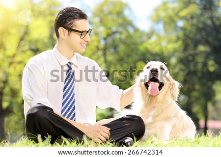 Smiling young businessman with his dog sitting on grass in a park. The dog is golden retriever