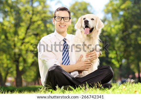 Young smiling man sitting on a green grass and hugging his labrador retriever dog in a park