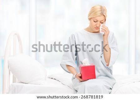 Sad woman in a hospital gown crying and wiping her tears seated on a hospital bed