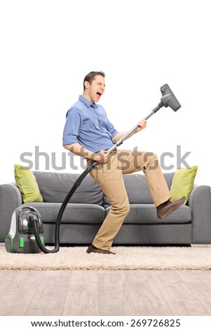 Excited young guy pretending to be playing guitar on the vacuum cleaner wand in front of a gray sofa isolated on white background