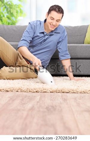 Vertical shot of a young man holding a handheld vacuum cleaner and cleaning a carpet at home