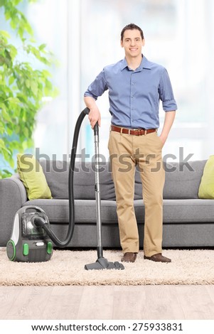 Full length portrait of a young man posing with a vacuum cleaner in front of a gray couch at home