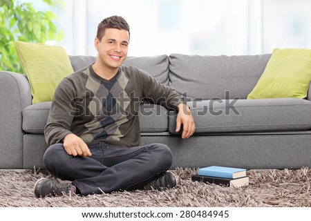 Young relaxed man sitting on the floor at home in front of a modern gray couch and looking at the camera