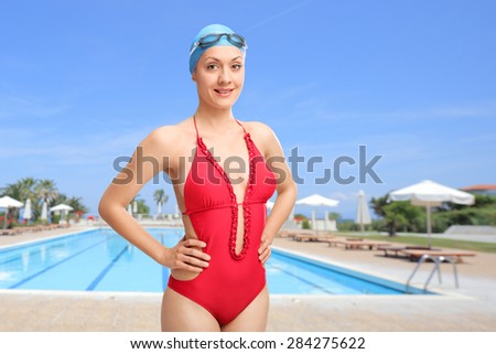 Young woman in red swimsuit wearing a blue swimming cap and goggles and posing in front of an open swimming pool
