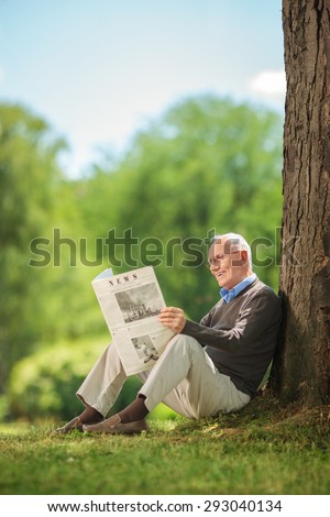 Senior man reading a newspaper in park and leaning against a tree. The newspaper is custom made the pictures are my copyright.