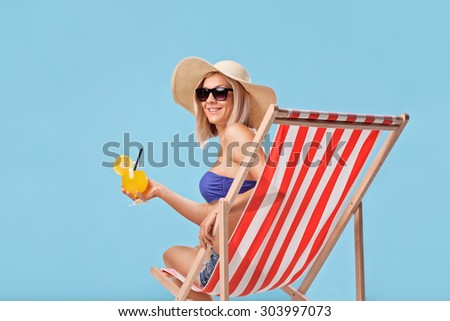 Young blond woman sitting in a sun lounger and holding an orange cocktail on blue background