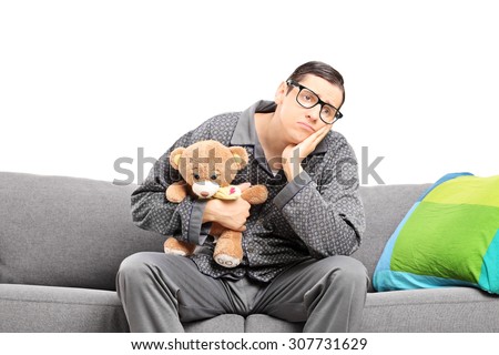 Sad man in pajamas holding a teddy bear seated on a sofa isolated on white