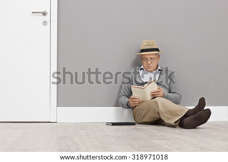 Senior gentleman reading a book seated on the floor and leaning against a wall next to a white door