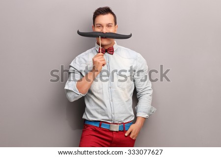 Cheerful young man holding a huge fake moustache below his nose and looking at the camera