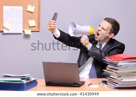 Angry businessman in an office, shouting on a megaphone, holding a mobile phone in the hand