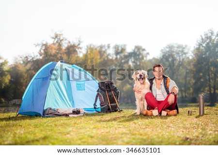 Young camper sitting in a meadow and hugging his pet dog next to a blue tent on a beautiful autumn day