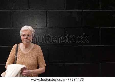 An old lonely woman is standing in front of a black wall
