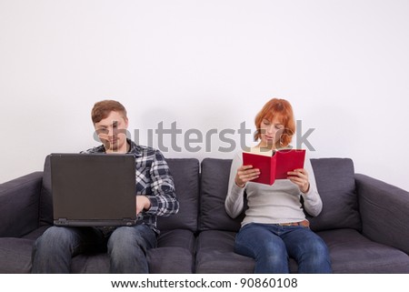 A young couple is sitting on the sofa. She is reading on he is working on his laptop.
