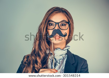 Close up portrait head shot beautiful, pretty young woman imitating a serious man. Attractive girl holding paper mustache isolated on gray green background. Positive human emotions, facial expressions