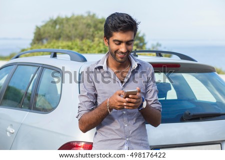 One young handsome Indian man smiling holding looking at mobile phone. Cheerful asian model outdoors background next to his car calling texting. People and technology concept