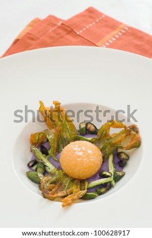 Fried egg\'s yolk over purple potatoes cream with asparagus and dehydrated zucchini\'s flowers. Served on a orange table cloth.