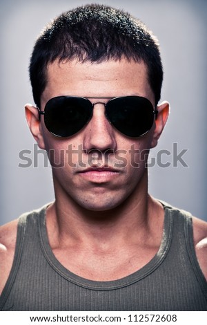 Stunning young man, with a very serious expression, appealing as the bad guy. The man is wearing sun glasses and some of his muscles are visible. A dramatic lighting has been used.