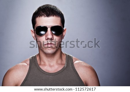Stunning young man, with a very serious expression, appealing as the bad guy. The man is wearing sun glasses and some of his muscles are visible. A dramatic lighting has been used.