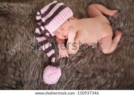 Newborn baby girl sleeping wearing brown and pink hat, on fur and white background.