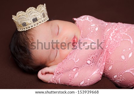 Adorable newborn baby sleeping wrapped on pink lace on brown cushion with a crown on the head.