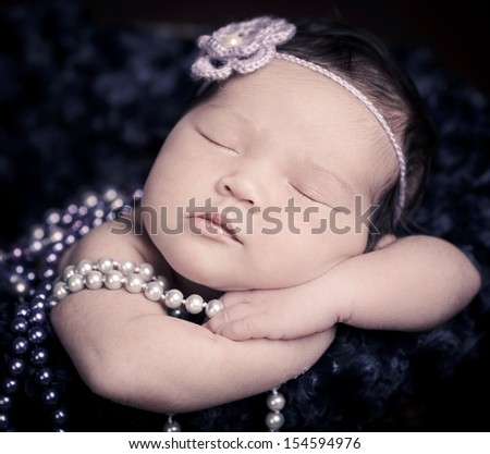 Newborn baby  sleeping at moon light inside a brown basket, resting on arms and elbows, on brown background. Wearing purple pearls and flower headband.