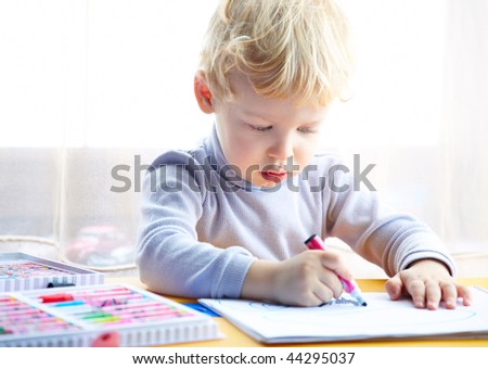 Little boy draws a colored pencil sitting at a table looking at the child a sheet of paper