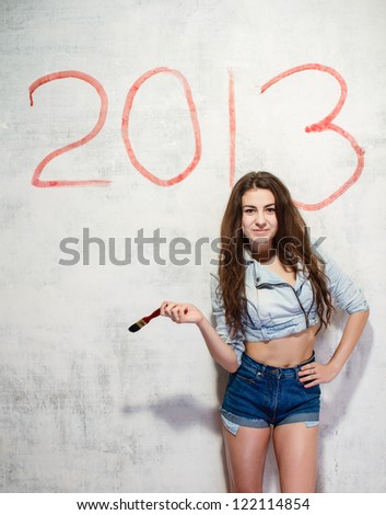 Girl in jeans shorts and jacket draws a Christmas (new year) decoration on old white wall with the red paint.
