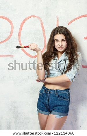 Girl in jeans shorts and jacket draws a Christmas (new year) decoration on old white wall with the red paint.