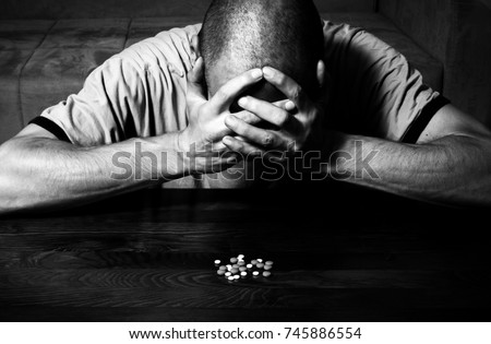 Depressed man suffering from suicidal depression want to commit suicide by taking strong medicament drugs and pills while he sitting on the floor of his dark room black and white image hard contrast