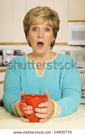 Senior woman, holding a coffee mug in her kitchen, with a look of shock on her face.