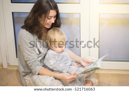 Portrait of happy young mother reading a book to her year-old baby boy with blue eyes sitting near a sunny window. Happy family lifestyle concept with solar flare