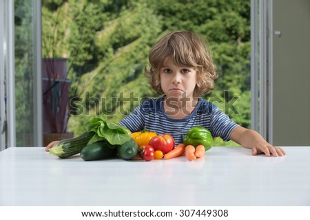 Cute little boy sitting at the table, unhappy with his vegetable meal, bad eating habits, nutrition and healthy eating concept