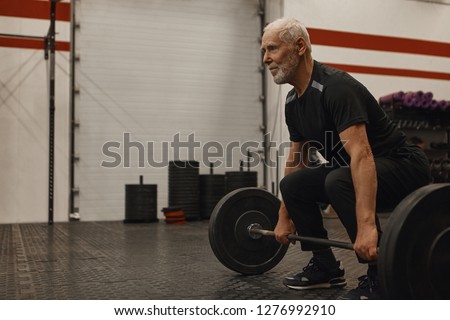 Stylish seventy year old male choosing healthy active lifestyle, training with weights indoors, lifting barbell, bulding muscular strong arms, having joufyl happy facial expression. Age and vitality