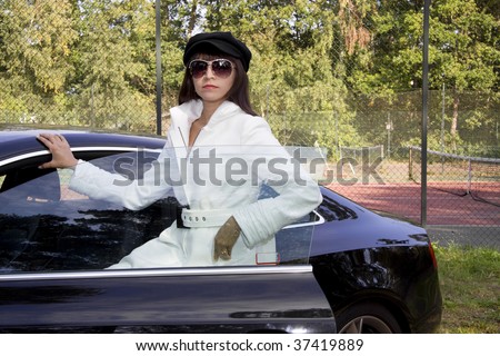 Fashion model wearing a white coat and a black beret hat standing outside a luxury car